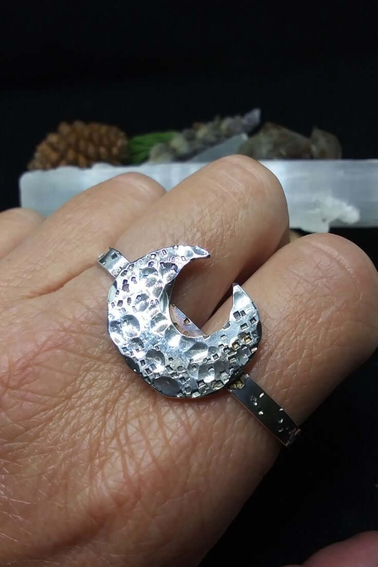 Highly polished silver double ring with a crescent moon motif that has hammered texture on both the crescent moon and band. Ring is worn on a hand with fingers folded down on a dark background with rocks and botanicals.