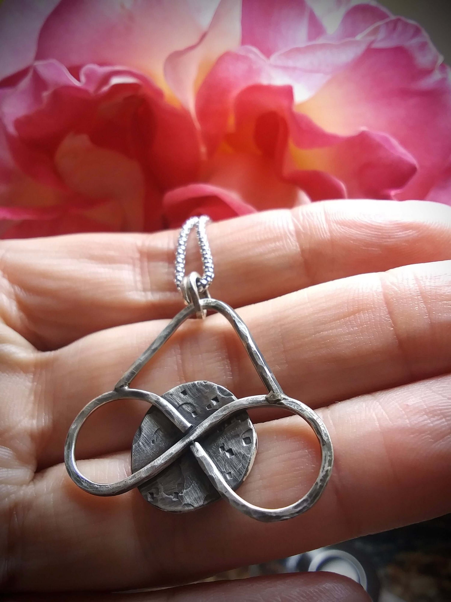 Silver pendant of a horizontal infinity sign overlaid n a textured full moon that is rested along the palm side of fingers in front of a large pink and yellowish rose.