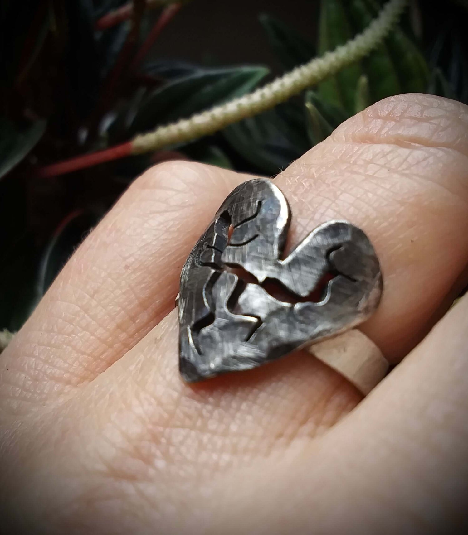 A dark silver asymmetrical heart ring that has pierced cracks and breaks worn on a folded finger in front of a botanical background.