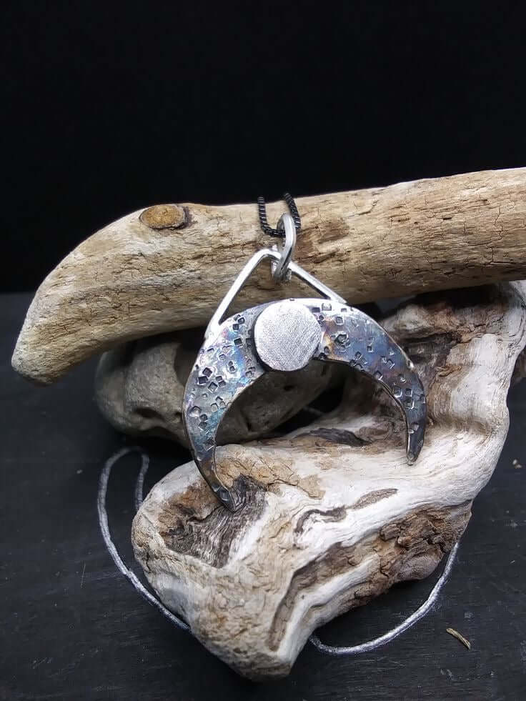 Silver crescent moon pendant that is textured with dark patina and rested amid pieces of driftwood on a dark background.