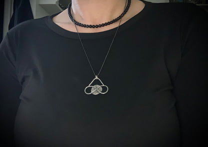  Medium sized silver pendant with dark patina that depicts a horizontal eternity symbol inlaid over a full moon on a necklace worn by a model in a black top.