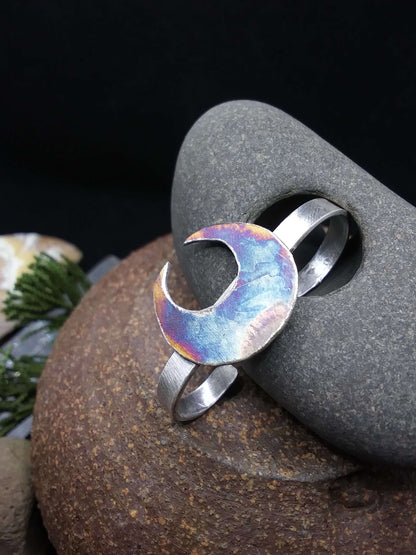 A silver double ring with a crescent moon that has unique patina, rested next to a hagstone.