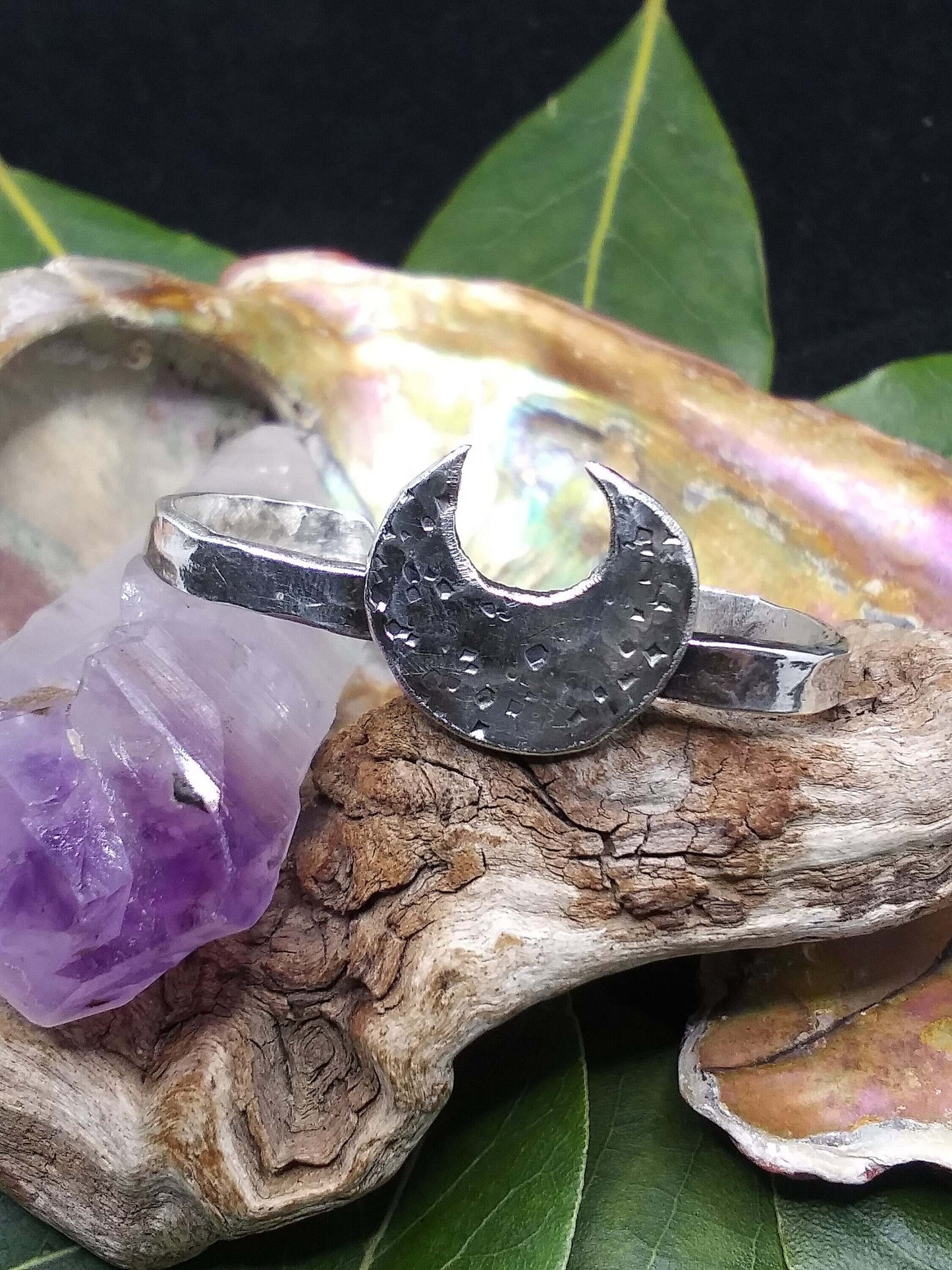 A silver double ring with a crescent moon that has hammered texturing rested on an abalone shell piece beside an amethyst crystal.