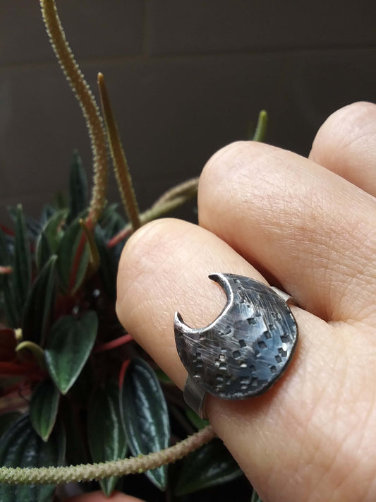 Dark silver crescent moon ring that has a big belly and hammered texturing, worn on a folded forefinger in front of dark colored botanicals.