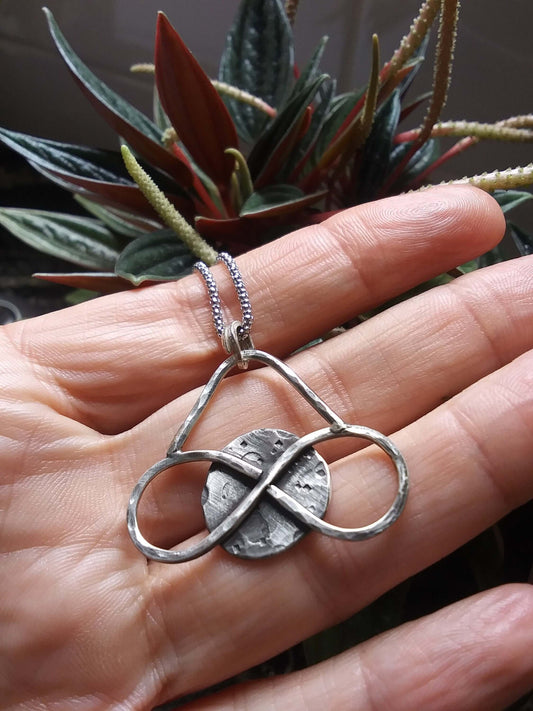 Medium sized silver pendant with dark patina that depicts a horizontal eternity symbol inlaid over a full moon. The pendant is rested on a palm in front of dark botanicals.