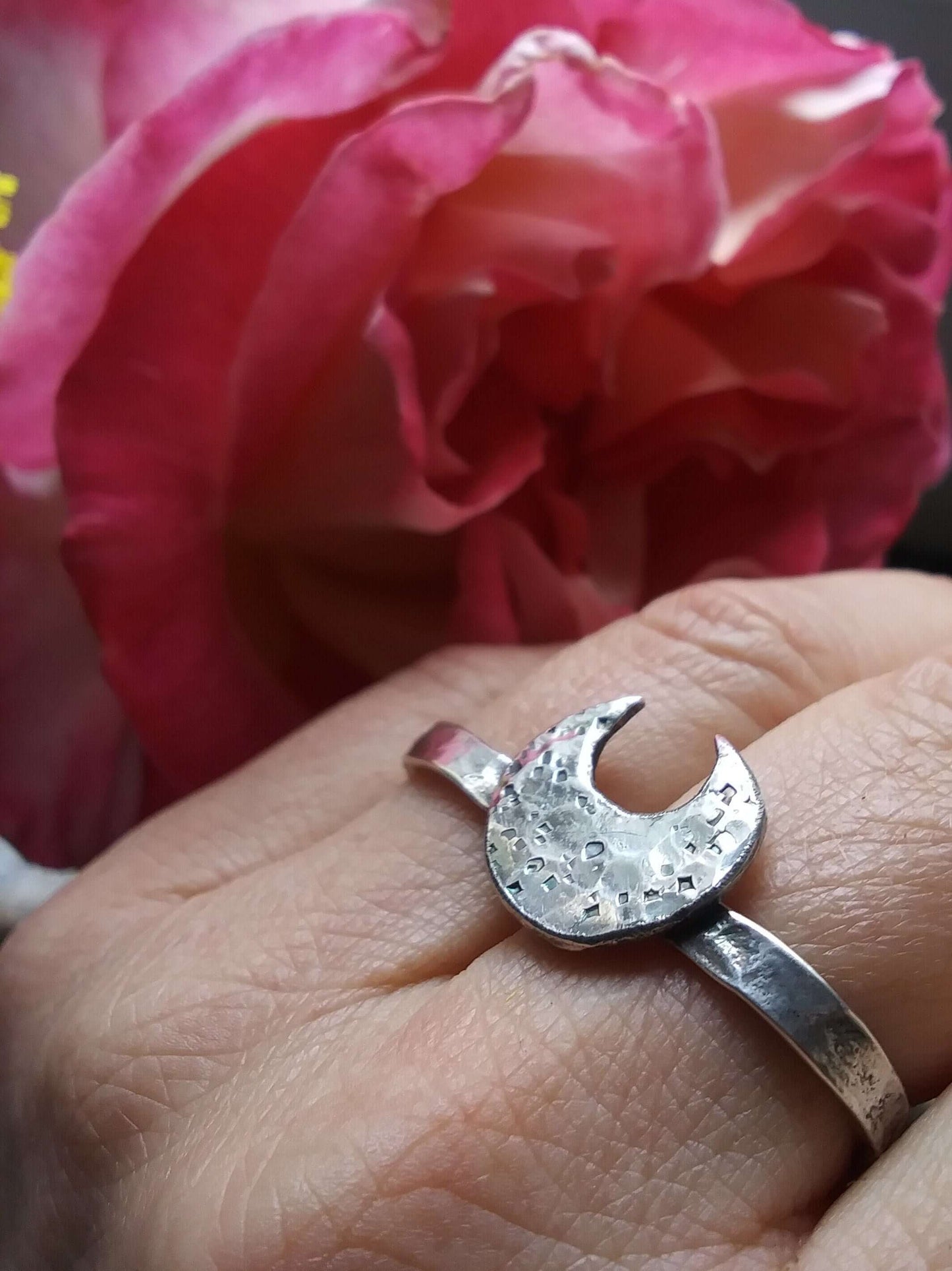 A silver double ring with a crescent moon that has hammered texturing on a hand with folded fingers in front of a large pink rose blossom.