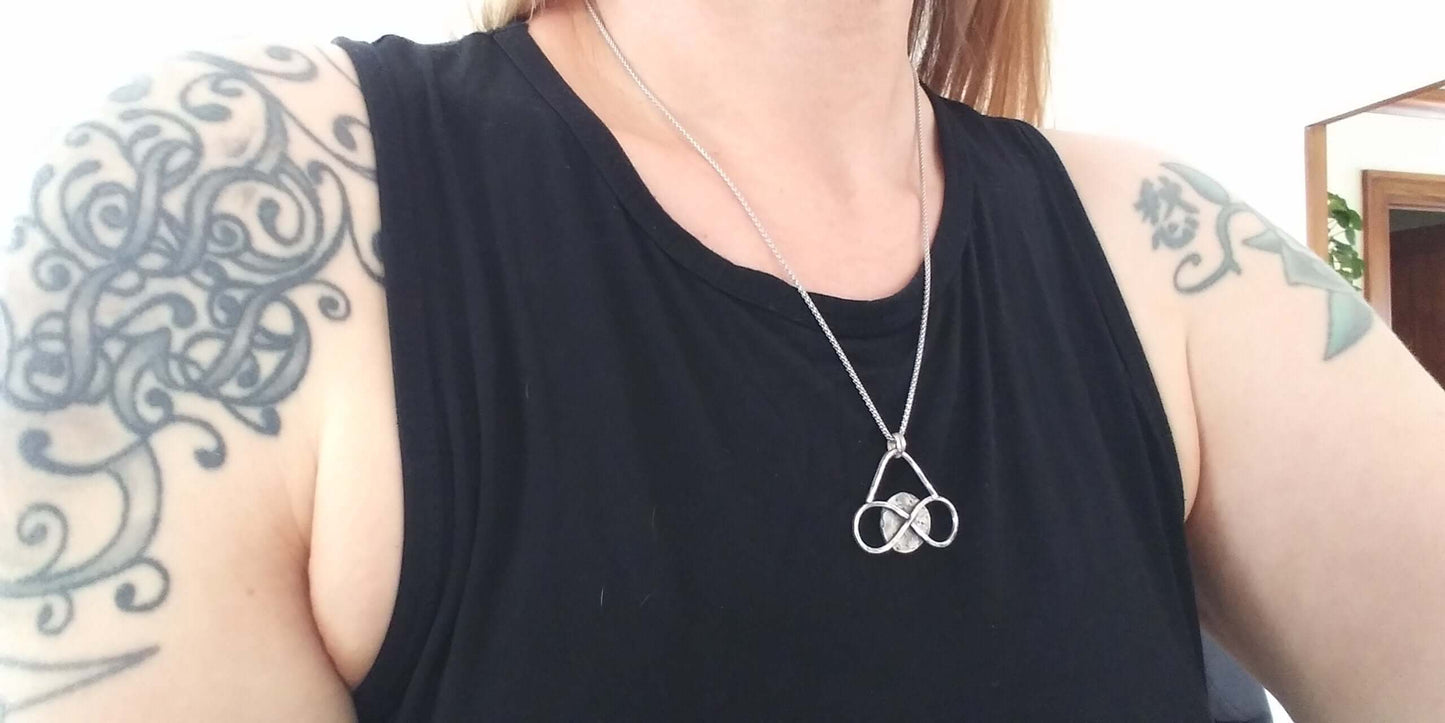  Medium sized silver pendant with dark patina that depicts a horizontal eternity symbol inlaid over a full moon on a necklace worn by a model in a sleeveless black top with shoulder tattoos.