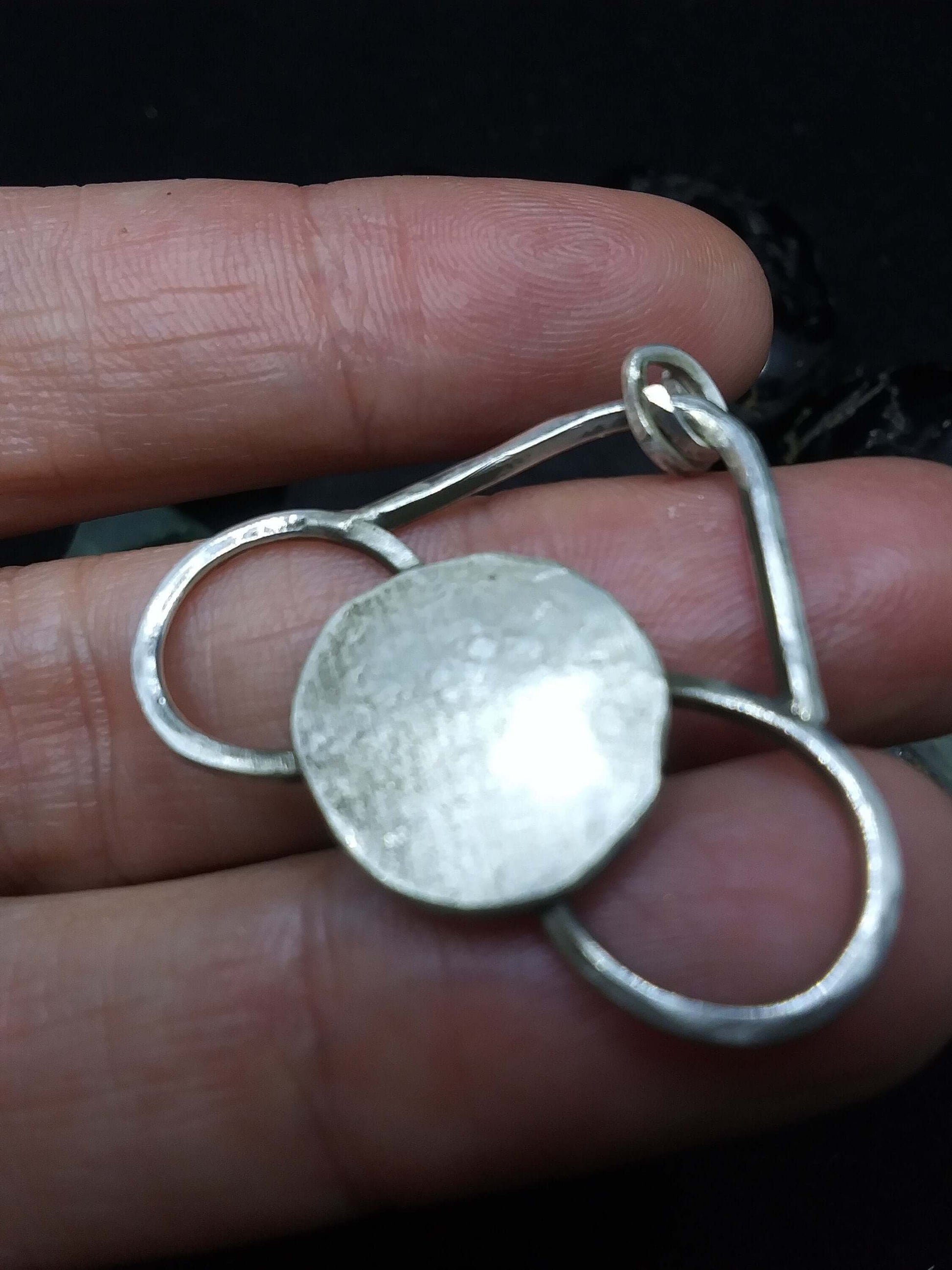  Back side of medium sized silver pendant with dark patina that depicts a horizontal eternity symbol inlaid over a full moon on palm side of fingers.