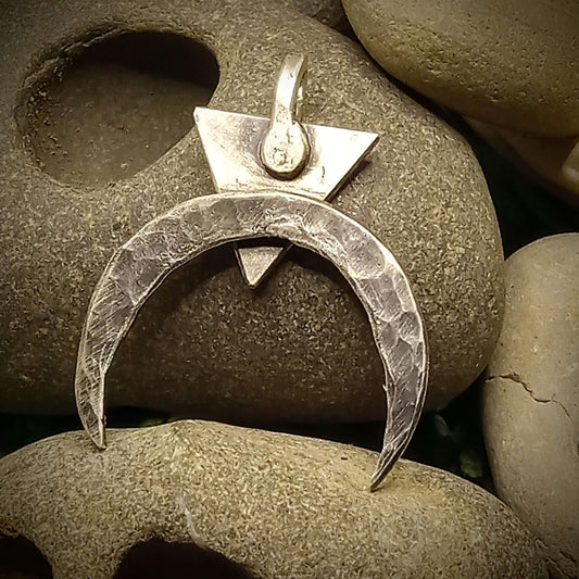A thin crescent moon pendant with a triangle behind it, both moon and triangle points are down. The pendant is rested on a hagstone.